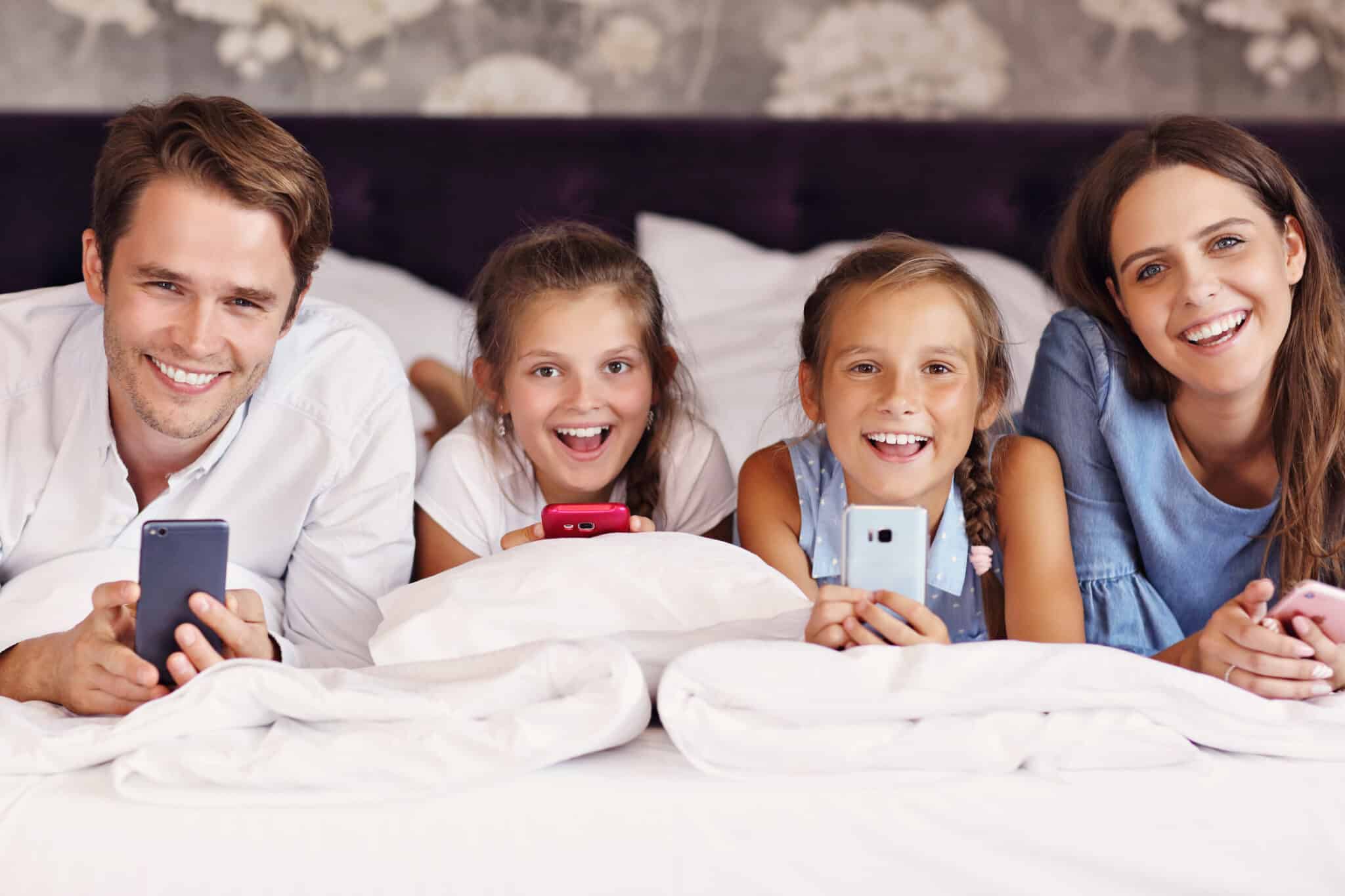 Familie mit zwei Mädchen und Eltern, die auf einem Bett in einem Hotelzimmer entspannen und Smartphones benutzen.