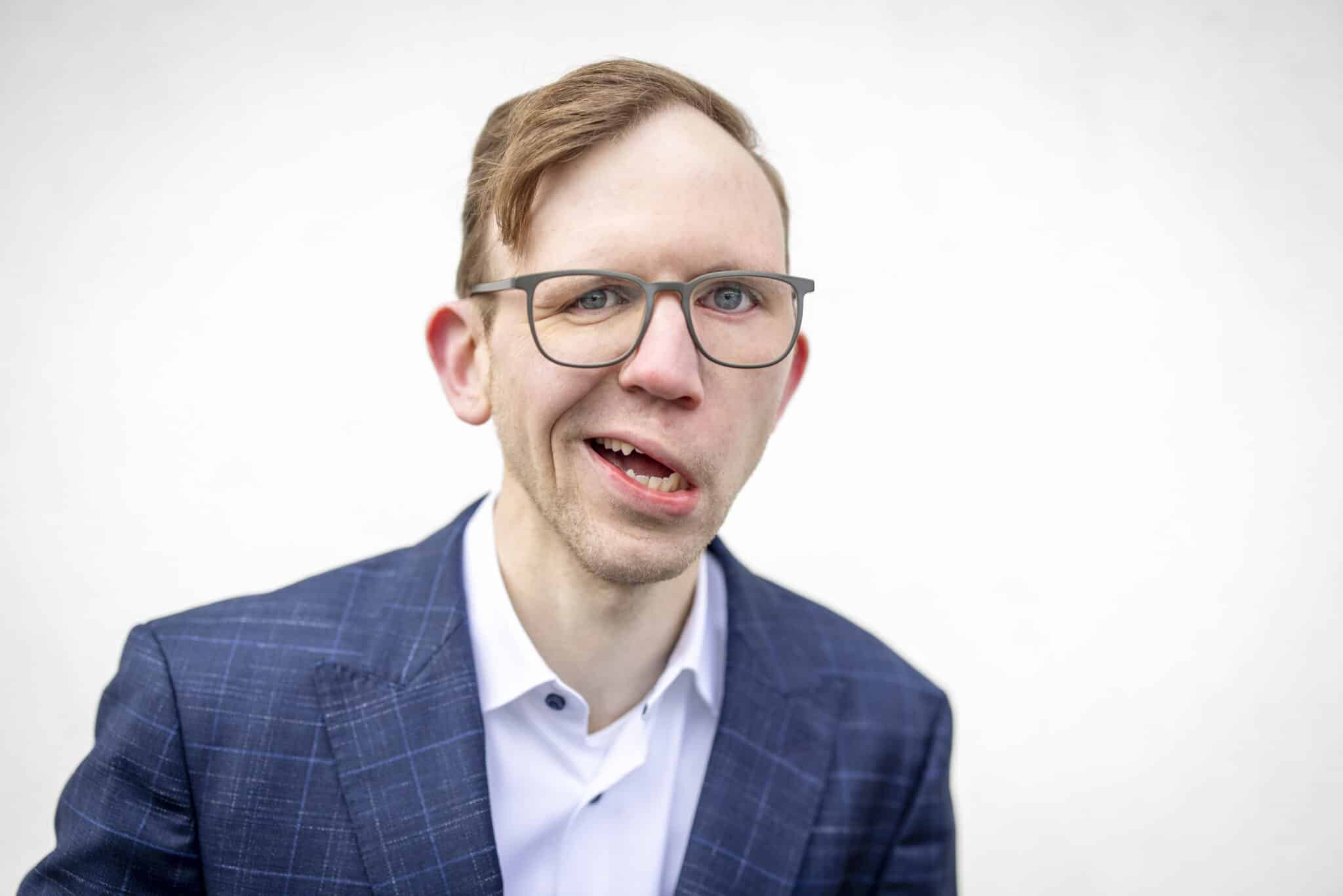 Marcel Friederich, eine Person mit hellbraunen Haaren und grauer Brille, trägt ein dunkelblaues Anzugjacke mit feinem Muster und ein weißes Hemd, lächelt leicht vor hellem Hintergrund.